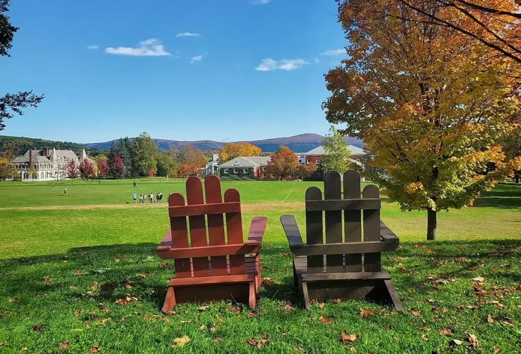 Two large Adirondack chairs on a grassy lawn at Middlebury College, looking out over a sports field towards historic campus buildings and mountains with fall foliage.