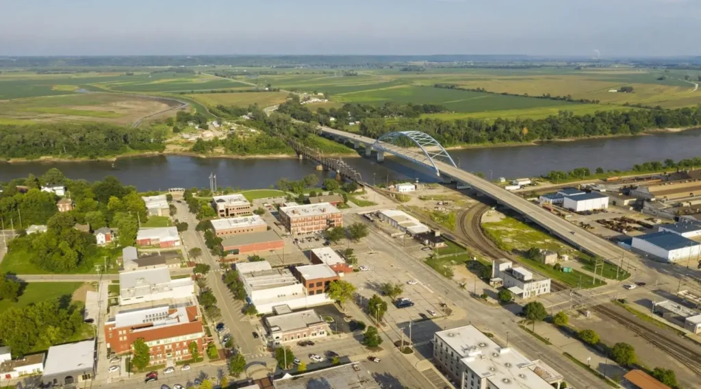 An aerial view of Atchison, Kansas, showing the historic downtown area, the Amelia Earhart Memorial Bridge crossing the Missouri River, and the surrounding green landscape.