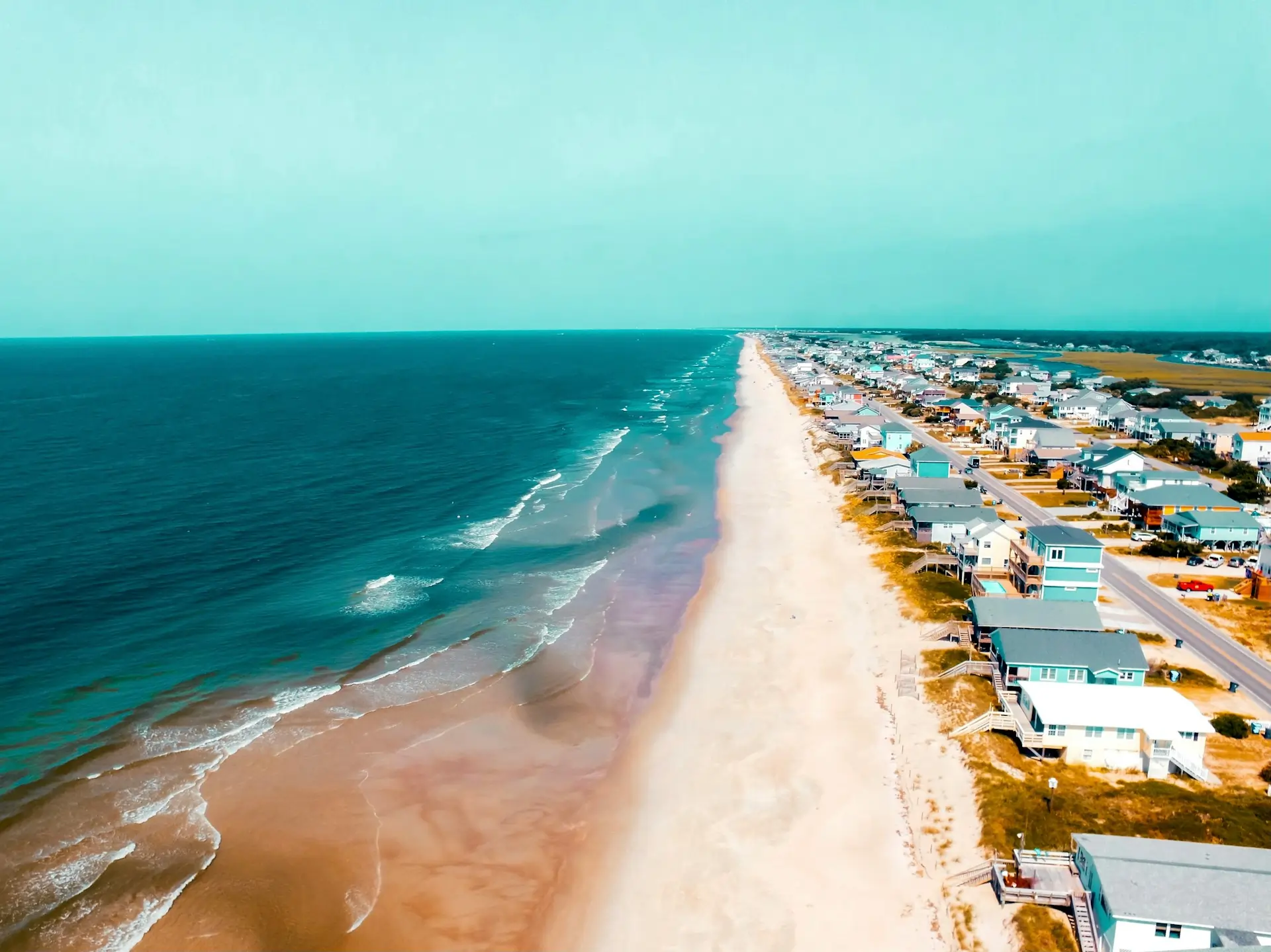 An aerial view of one of the many beautiful North Carolina beaches, showing a long stretch of sand, ocean waves, and beachfront houses.