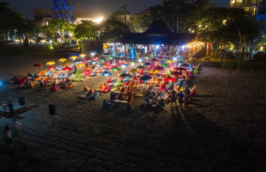 A vibrant beach club in Bali at night, one of the top cities in Asia for nightlife, with people relaxing on colorful beanbags under umbrellas.