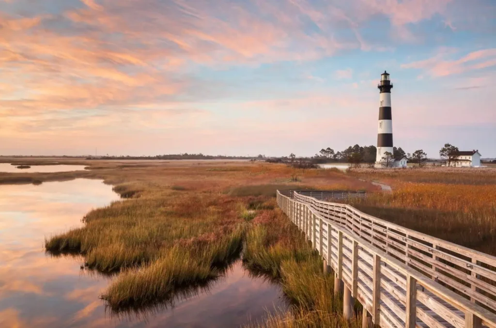 The Bodie Island Lighthouse on the Outer Banks, an iconic landmark of the North Carolina beaches, seen at sunset with a wooden boardwalk leading across a marsh.
