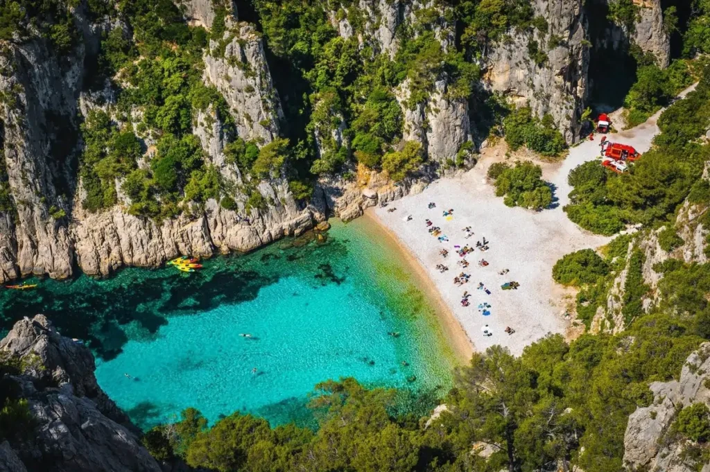 An aerial view of the secluded Calanque d'En-Vau near Cassis, France, one of the best summer destinations in Europe, showing its white pebble beach and turquoise water surrounded by cliffs.