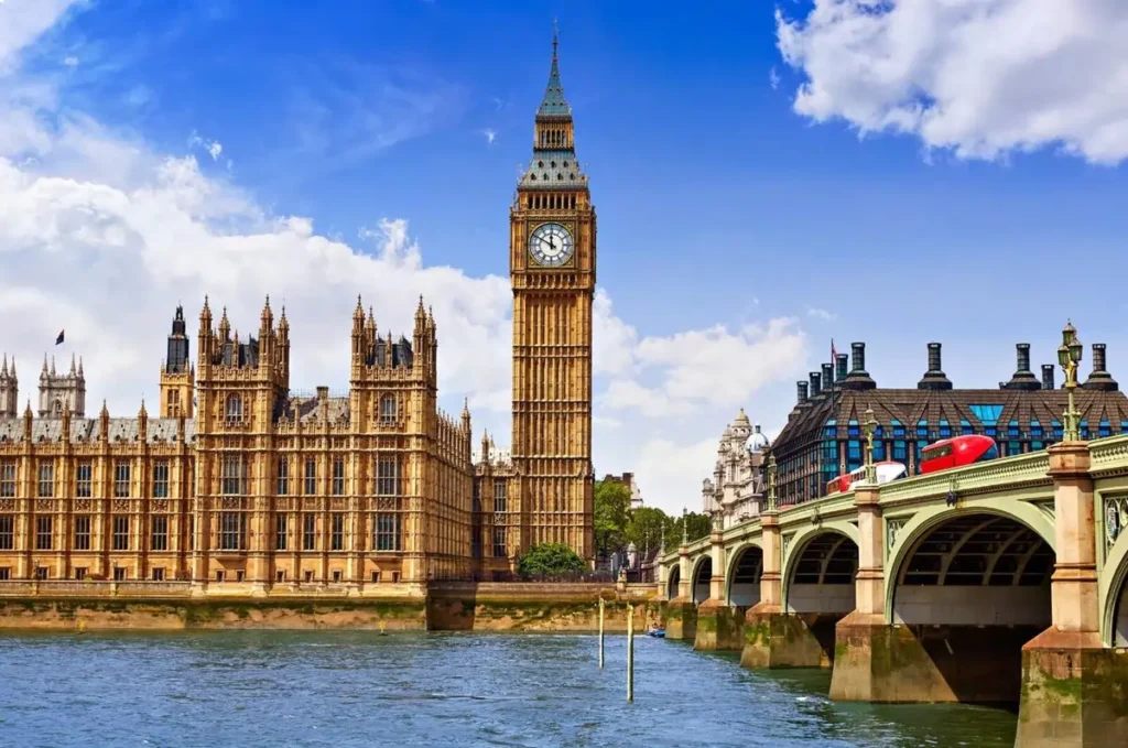 The Houses of Parliament and Big Ben on the River Thames in London, with a red double-decker bus crossing Westminster Bridge.