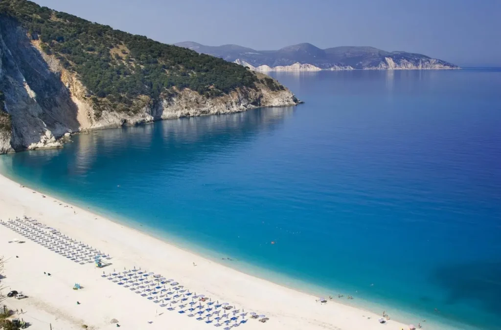 An aerial view of the world-famous Myrtos Beach in Kefalonia, Greece, one of the best summer destinations in Europe, showing its white pebble shore and electric-blue water.