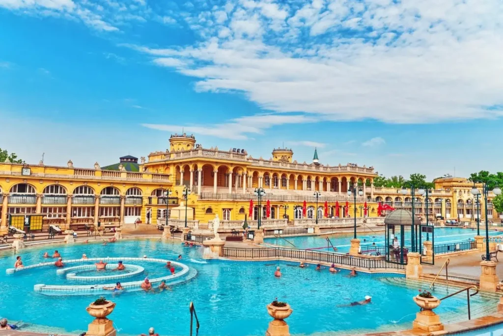 People relaxing in the large outdoor pools of the Széchenyi Thermal Bath in Budapest, with the grand, yellow neo-baroque palace in the background.