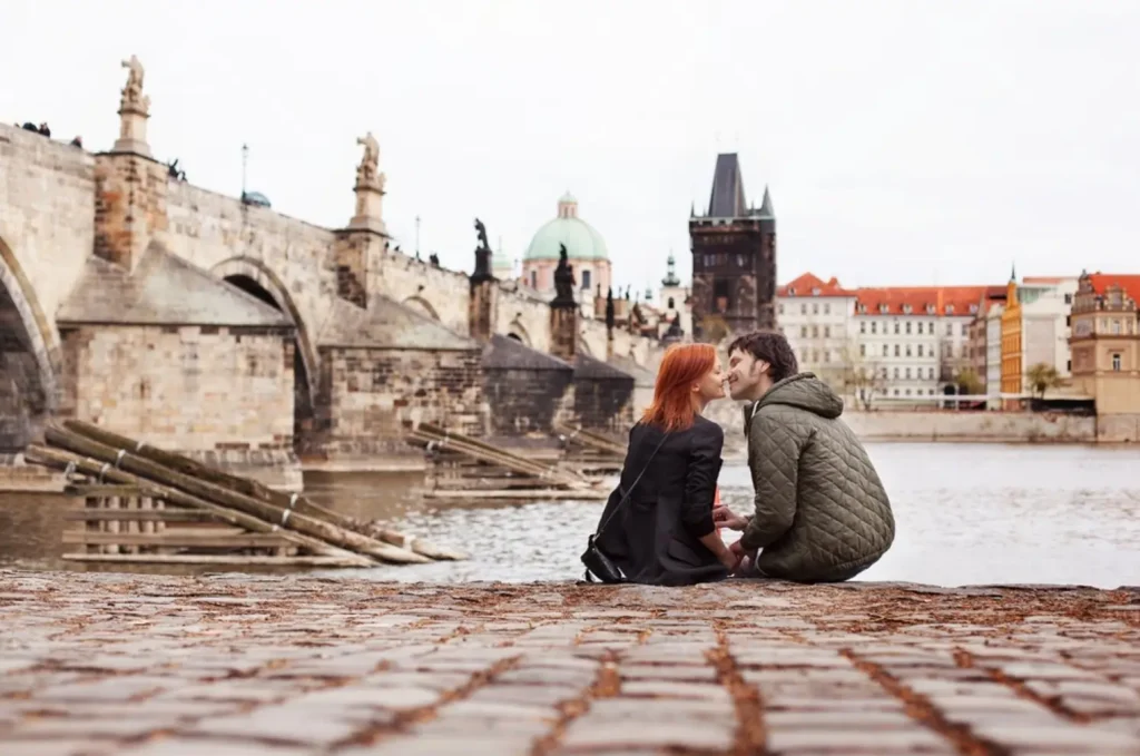A romantic couple about to kiss on the cobblestone banks of the Vltava River in Prague, with the historic Charles Bridge in the background, making it one of the best honeymoon destinations in Europe.