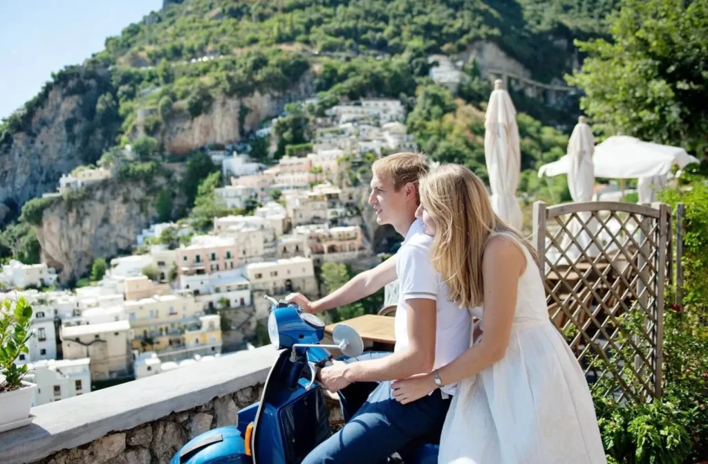 A honeymoon couple on a blue scooter overlooking the cliffside village of Positano on the Amalfi Coast, one of the best honeymoon destinations in Europe.