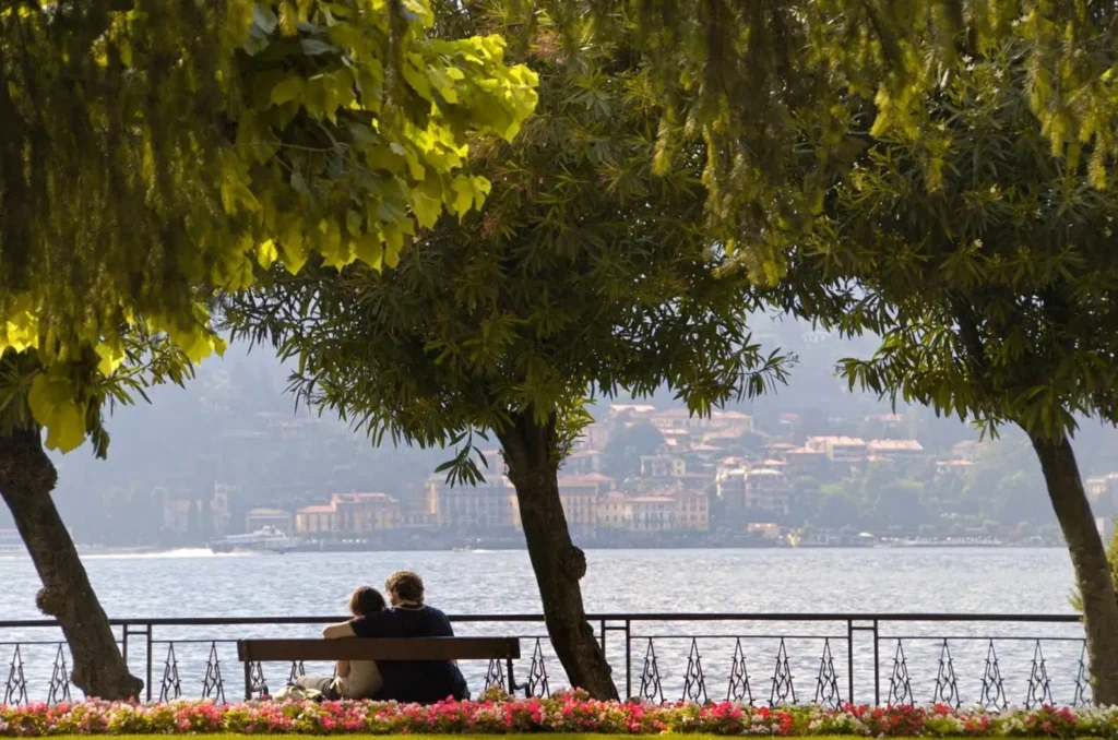 A couple on their honeymoon sitting on a bench overlooking Lake Como, Italy, one of the best honeymoon destinations in Europe, framed by lush trees and flowers.