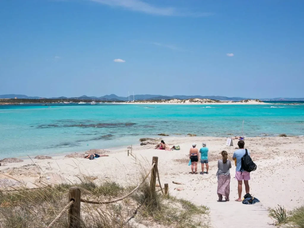 People on a wooden boardwalk leading to the white sand and brilliant turquoise water of Ses Illetes beach in Formentera, one of the best summer destinations in Europe.