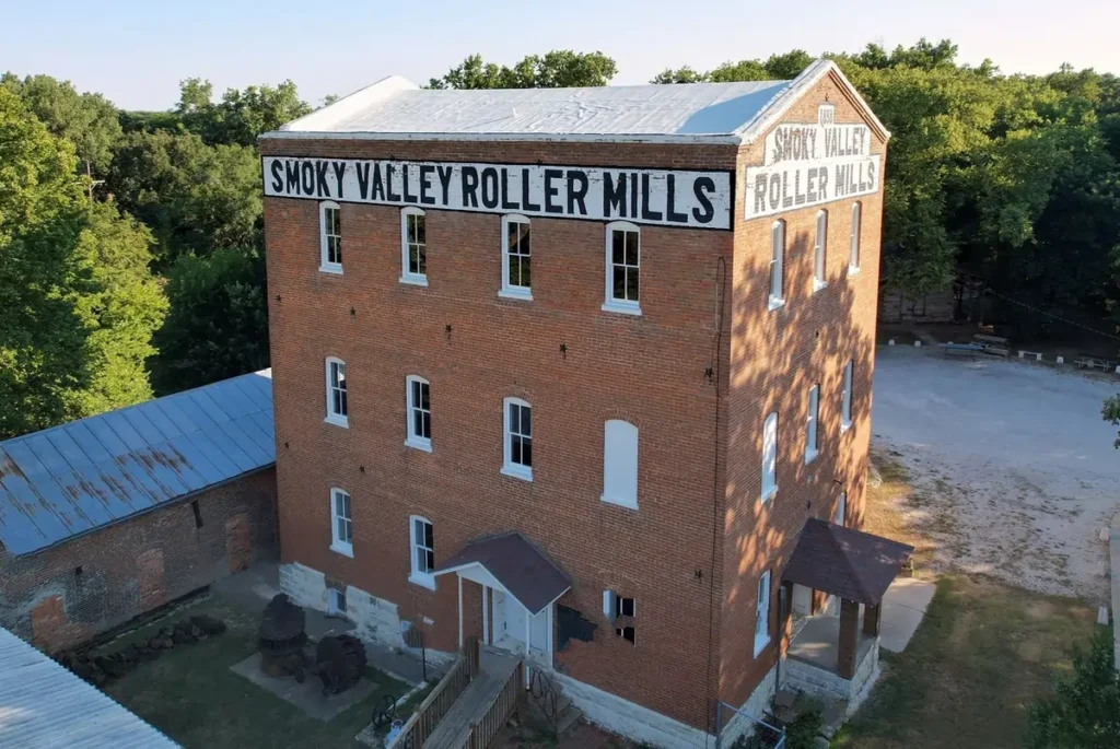 An aerial view of the historic, multi-story brick Smoky Valley Roller Mills building in Lindsborg, Kansas, surrounded by green trees on a sunny day.