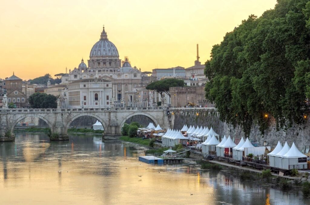 A view of St. Peter's Basilica and the Ponte Sant'Angelo bridge over the Tiber River in Rome at sunset, with a market along the riverbank.