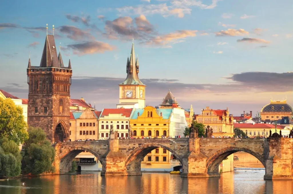 A panoramic view of the historic Charles Bridge in Prague at sunset, with the Old Town Bridge Tower and colorful buildings reflecting in the Vltava River.