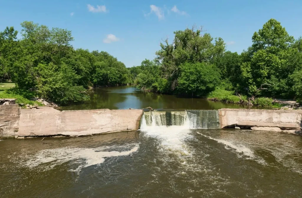 The namesake waterfall on the Cottonwood River in Cottonwood Falls, Kansas, with water flowing over a historic dam surrounded by lush green trees.