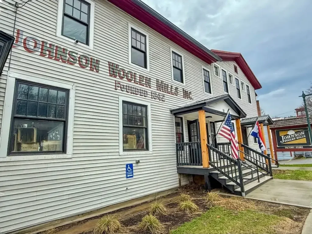 The historic, white clapboard building of Johnson Woolen Mills in Johnson, Vermont, with its name painted on the side and an American flag at the entrance.