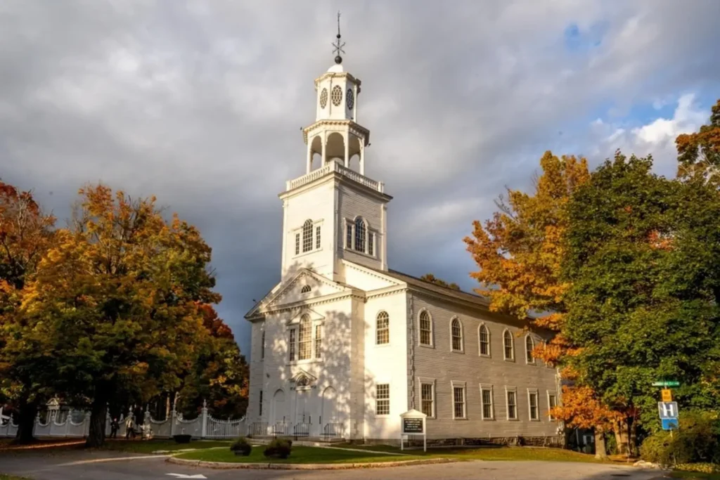 The historic, white Old First Congregational Church in Bennington, Vermont, framed by trees with beautiful autumn foliage under a dramatic sky.