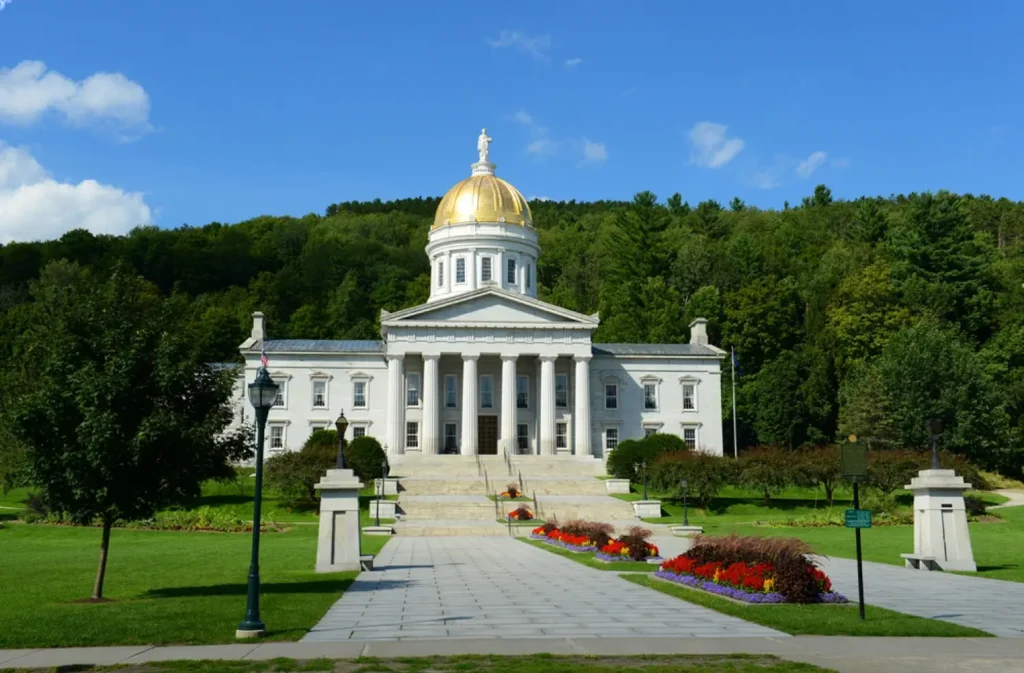 The Vermont State House in Montpelier, a white neoclassical building with a large gold dome, set against a backdrop of a lush green hill.