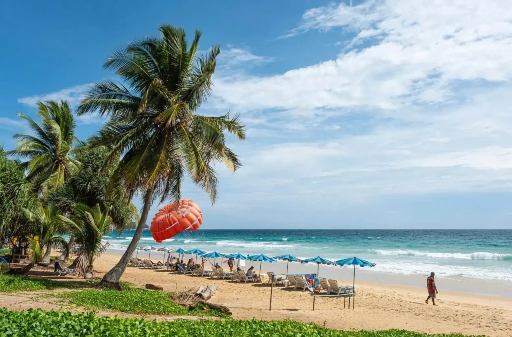 A beautiful beach in Phuket, one of the best cities in Asia for a tropical honeymoon, with palm trees, beach chairs, and a parasail.