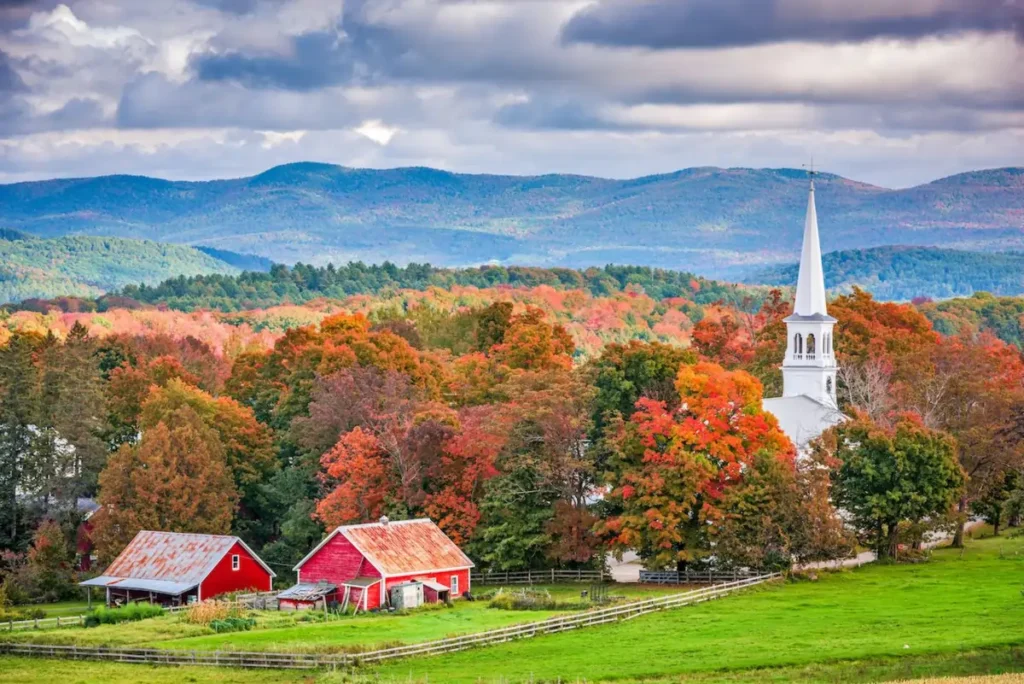Vermont fall landscape with vibrant foliage, rustic barns, and a white church steeple against rolling hills 