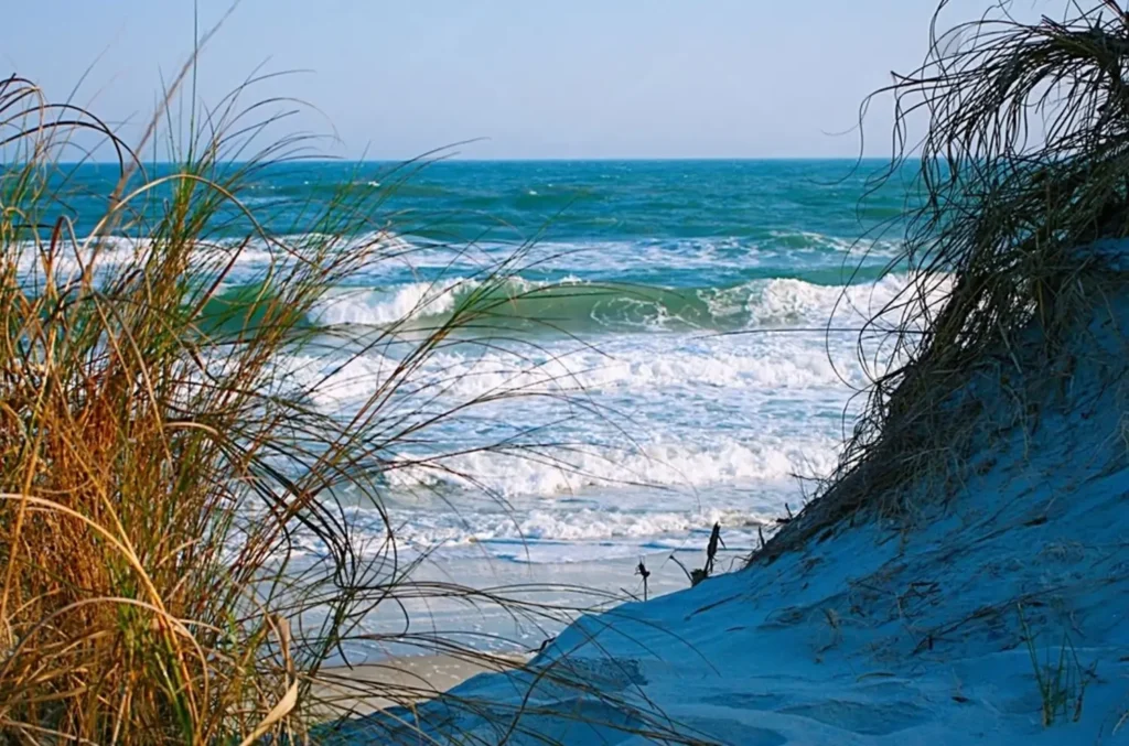 A view of the ocean waves from behind a sand dune with sea grass at Hammocks Beach State Park, one of the most natural North Carolina beaches.