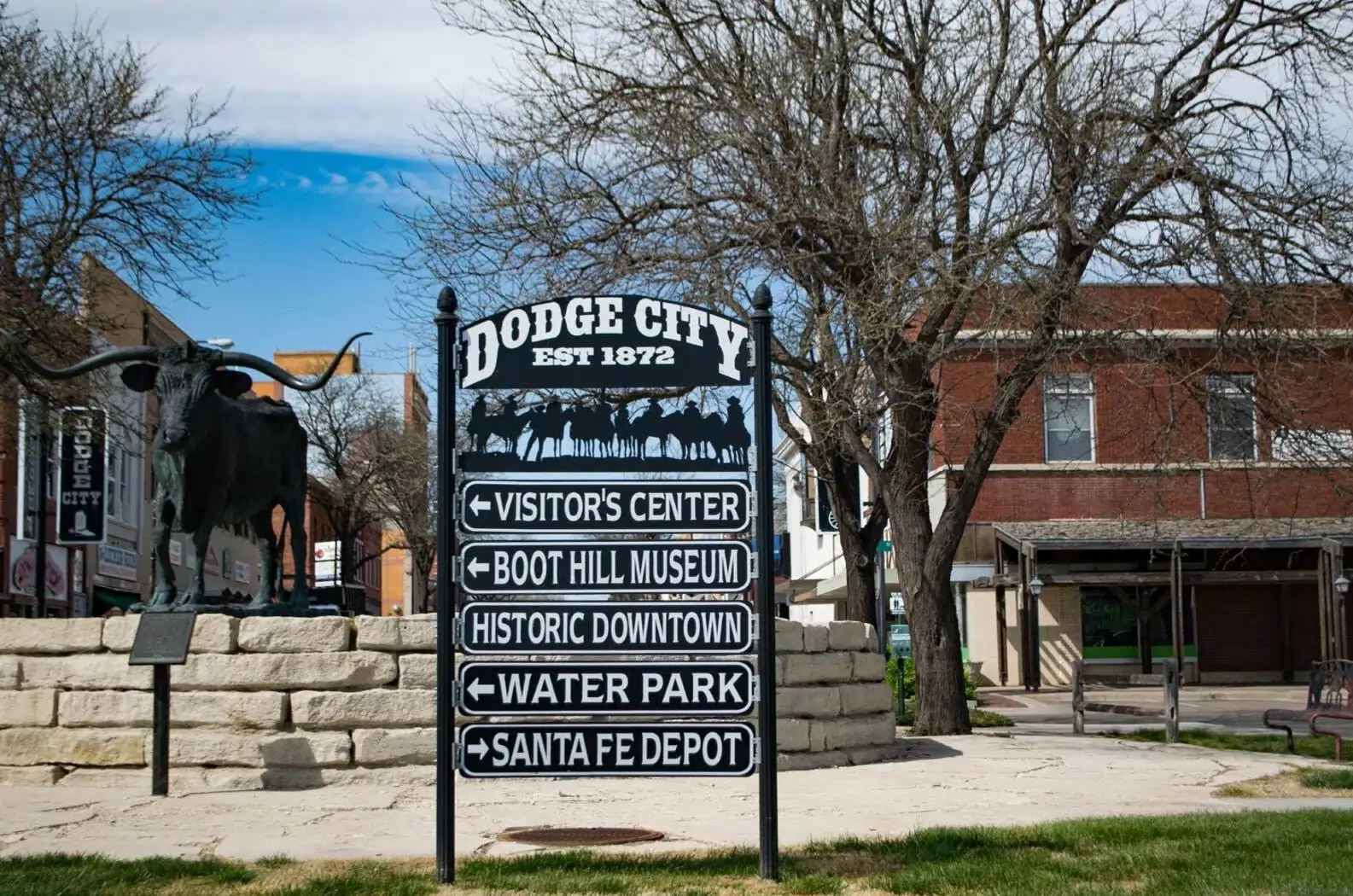 A directional sign in historic downtown Dodge City, Kansas, pointing to family vacation attractions like the Boot Hill Museum and Visitor's Center, with a longhorn cattle statue in the background.