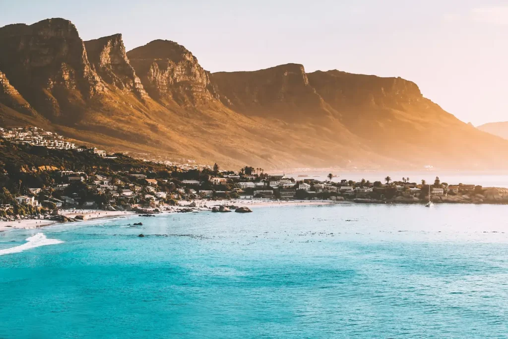 A scenic view of a beach in Cape Town, one of the best cities in South Africa for a safari combo trip, with mountains meeting the turquoise ocean at sunset.