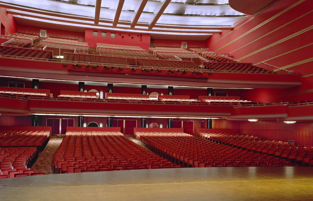 The grand interior of the Kansas City Music Hall, showing the empty red seats of the orchestra and balcony levels from the stage's perspective.