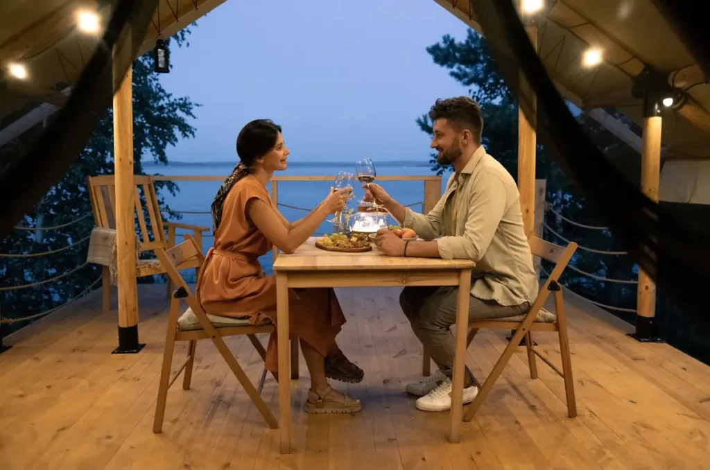 A couple enjoying a romantic waterfront dinner, toasting with wine glasses on a private deck overlooking the ocean at dusk.