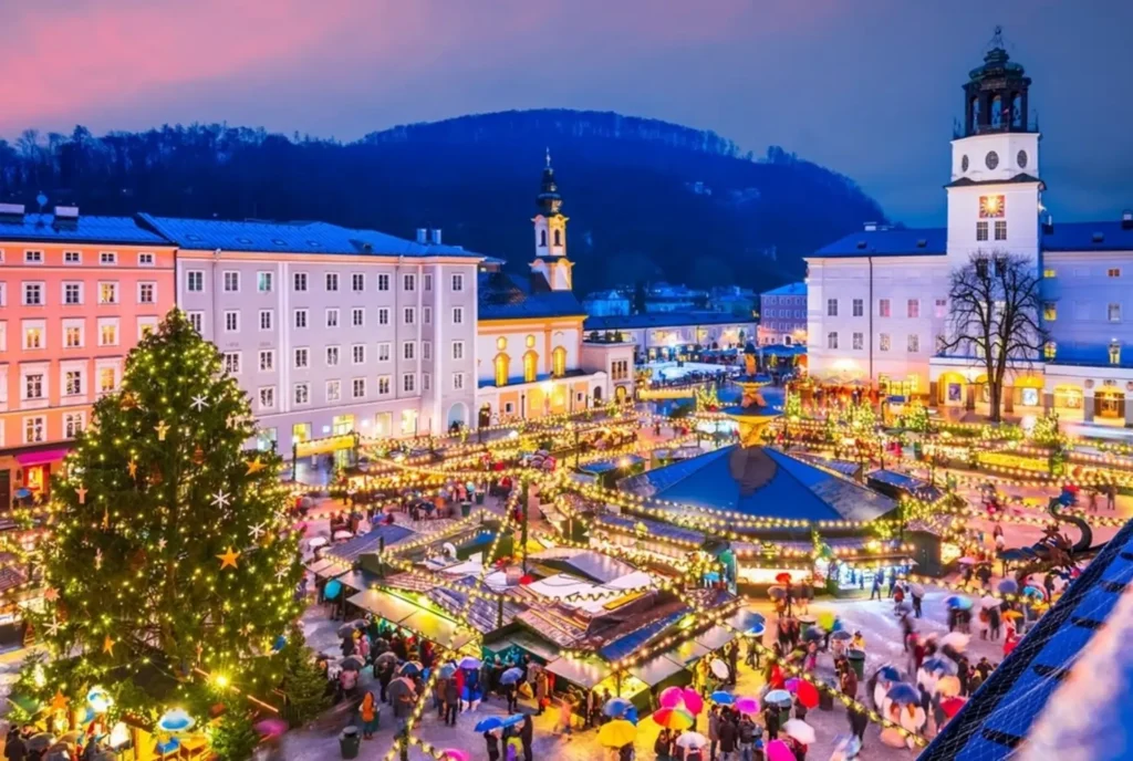 An aerial view of the Salzburg Christmas Market at dusk, one of the best Christmas markets in Europe, with a large Christmas tree and numerous illuminated stalls.