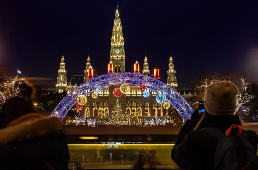 A visitor taking a photo of the Vienna Christmas Market at night, with the illuminated City Hall (Rathaus) and a festive archway in the background.